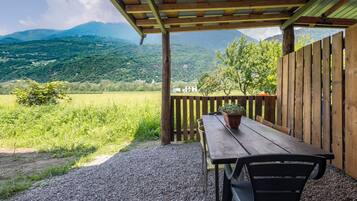 A covered outdoor seating area with a table and chairs, a potted plant, and a view of a green landscape with mountains in the background.