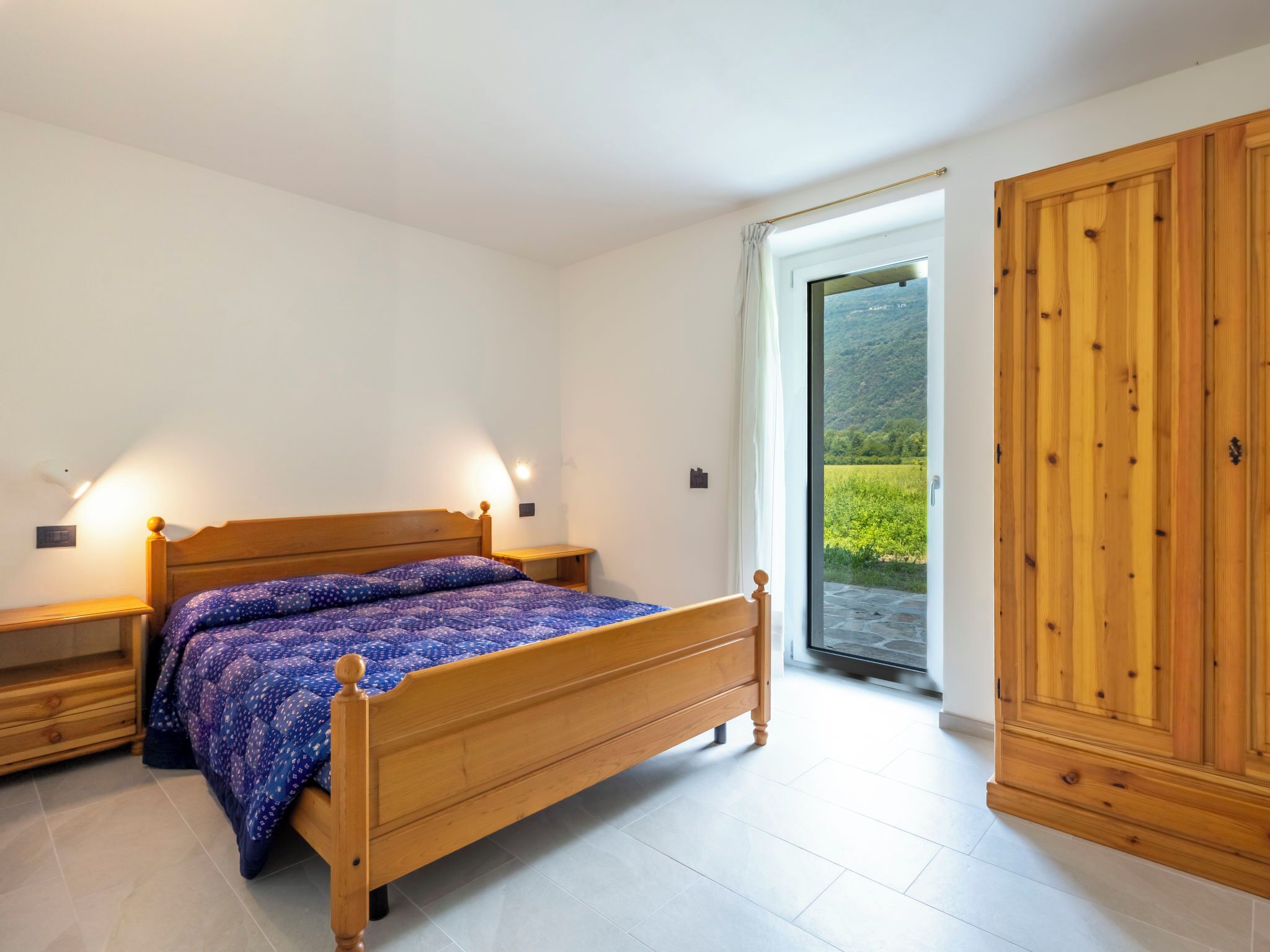Une chambre à coucher avec un lit en bois, des tables de chevet, une armoire et une vue sur un paysage verdoyant, visible à travers une porte vitrée.