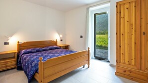 Quarto com cama de madeira, mesas de cabeceira, um guarda-roupa e vista para uma paisagem verde através de uma porta de vidro.