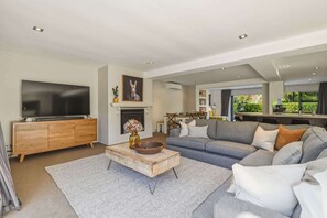A modern living room with a flat-screen TV, wooden cabinet, grey sofa, and a fireplace. - Lake Ōkareka - Spa Pool & Log Fire, Sleeps 12 (Rotorua)