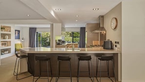 A modern kitchen with a central island, bar stools, a clock, and a view of greenery outside.