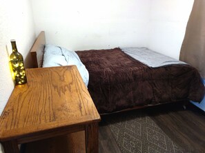 Ein kleiner Raum mit einem Holztisch, einer Weinflasche, einem braunen Bett mit Decke und einem gemusterten Teppich.