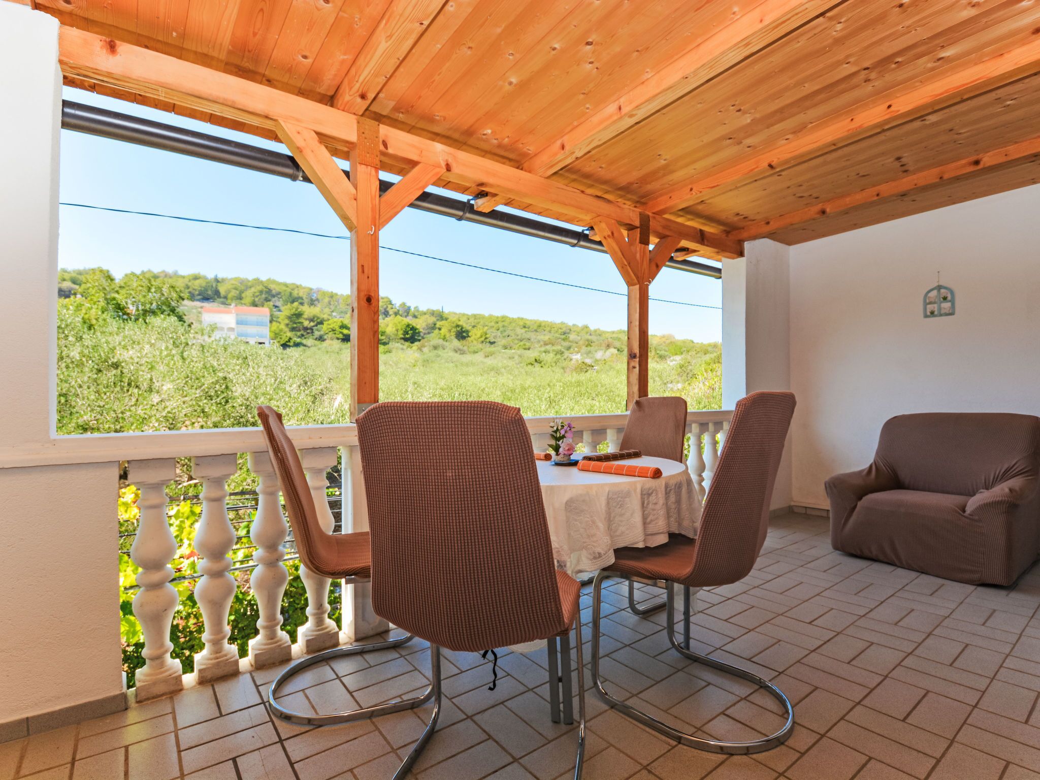 Une terrasse couverte avec une table et des chaises, une vue sur la verdure et les bâtiments, et un mur blanc agrémenté d’un petit objet décoratif.