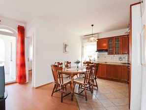 A dining area with a wooden table and chairs, a kitchen with wooden cabinets, and a window with curtains. - Apartment Jako by Interhome (Trogir/Slatine)