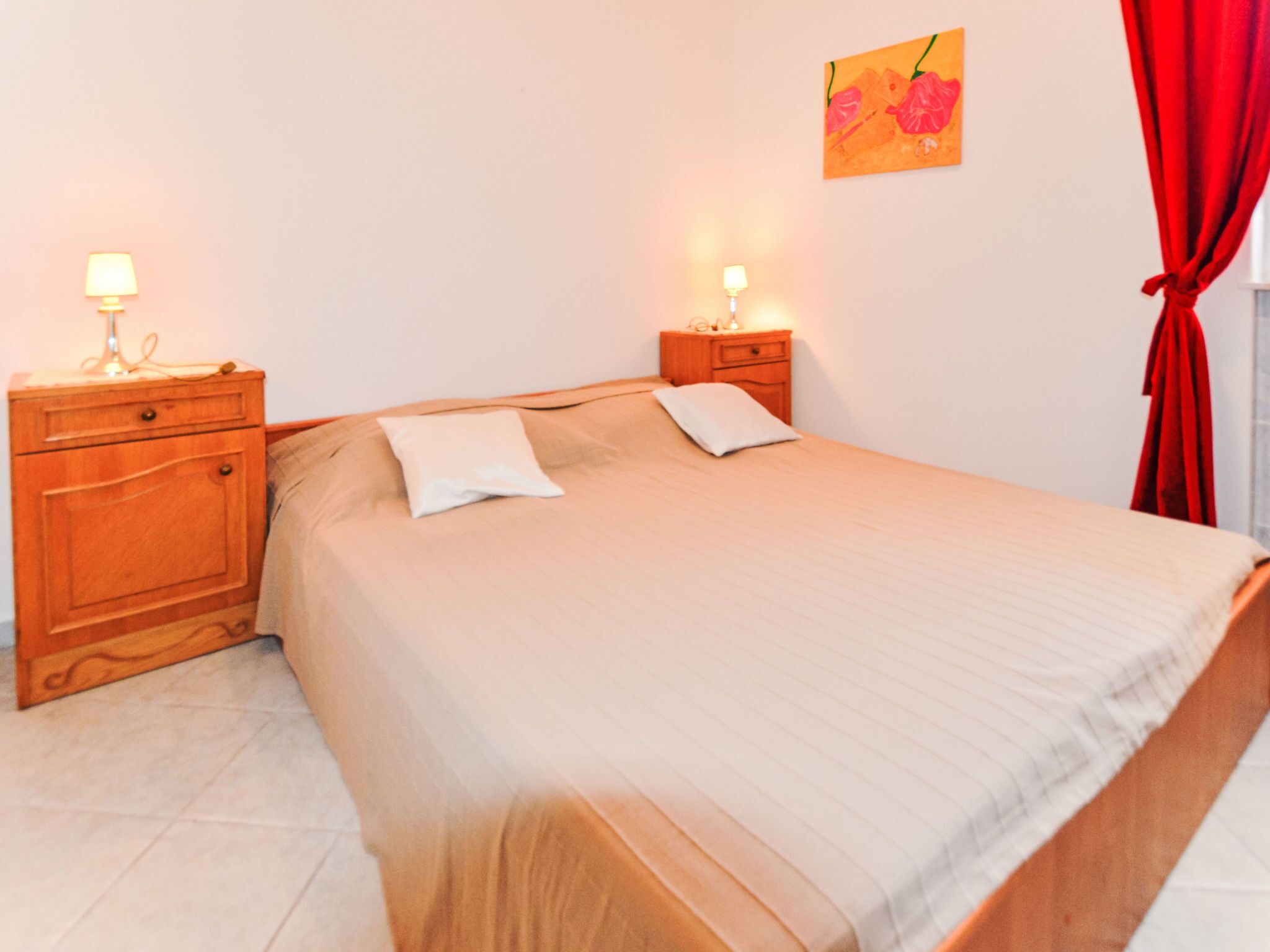 A bedroom with a bed, two bedside tables, a lamp on each table, a red curtain, and a painting on the wall.