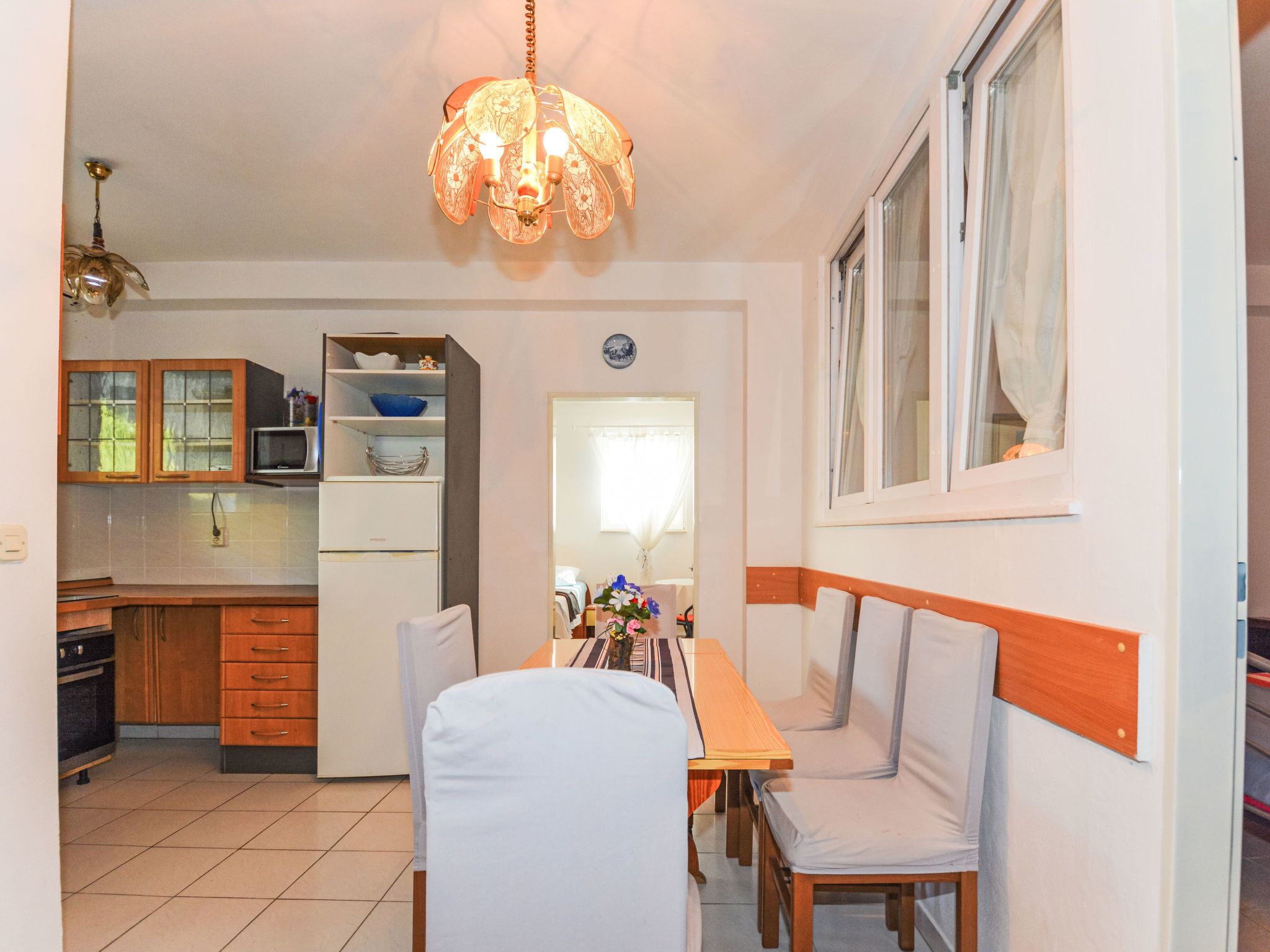 A compact kitchen with a microwave, refrigerator, and wooden cabinets. There is a dining area with a table and chairs, and a window with curtains.