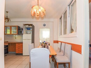 A compact kitchen with a microwave, refrigerator, and wooden cabinets. There is a dining area with a table and chairs, and a window with curtains. - Apartment Jako by Interhome (Trogir/Slatine)
