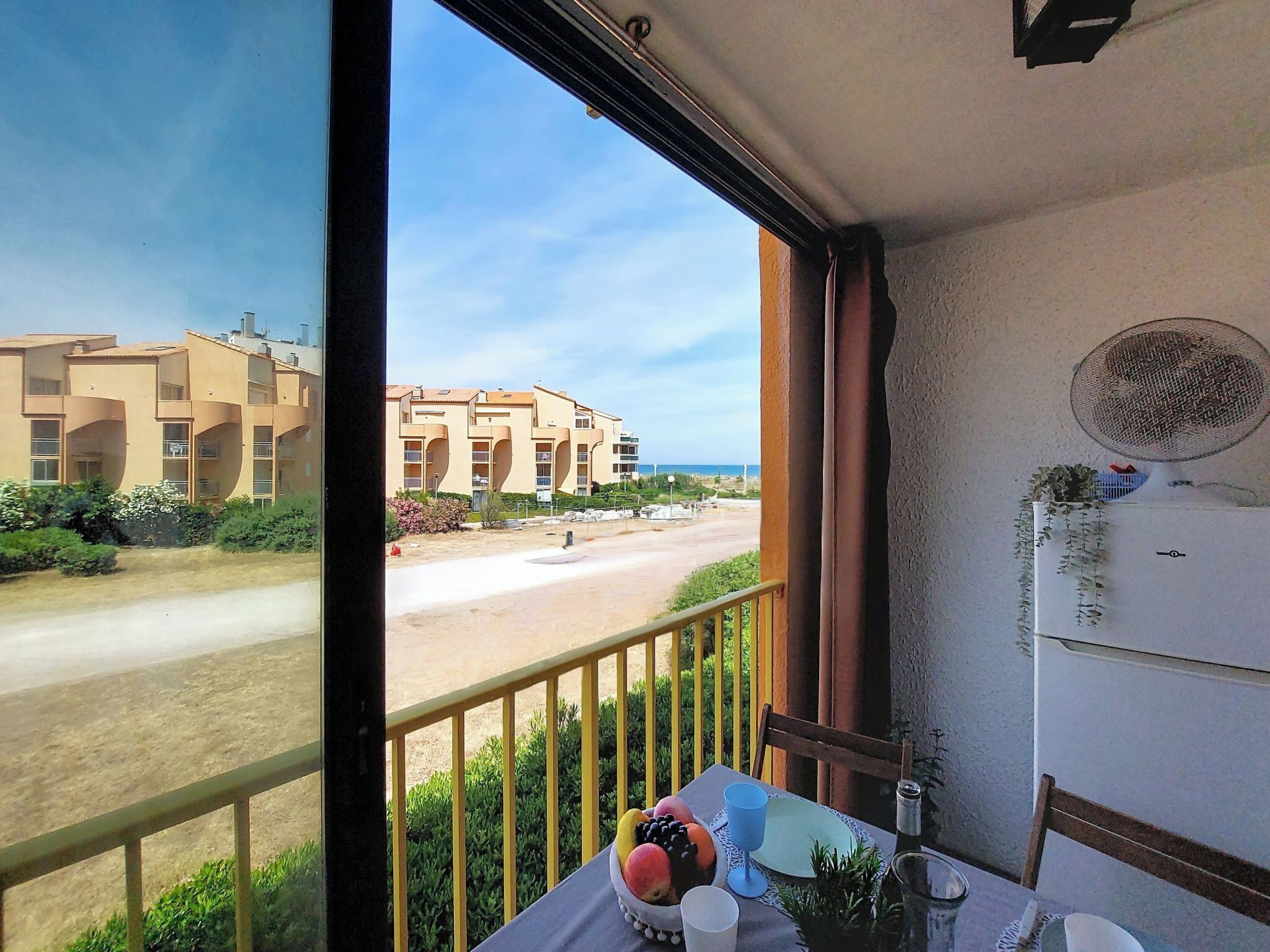 Ein Balkon mit Blick auf Strand und Gebäude, ein Tisch mit Früchten und einem Glas, sowie ein an der Wand befestigter Ventilator.