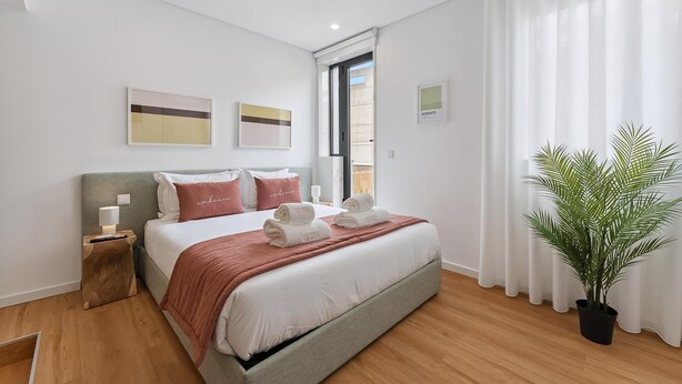 Una habitación de hotel moderna con una cama grande, una mesita de noche con lámpara, una planta en maceta y una ventana con cortinas.