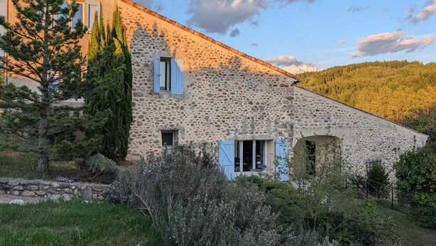 Un edificio de piedra con techo inclinado, una pequeña ventana con contraventanas y un muro de piedra.