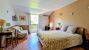 Un dormitorio con cama, escritorio con silla, una mesita, lámpara, un cuadro en la pared y balcón.
