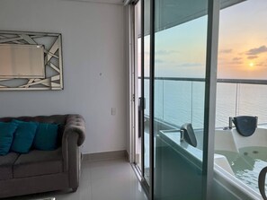 Ein Balkon mit Meerblick bei Sonnenuntergang, eine Sitzecke mit türkisfarbenen Kissen und ein modernes, abstraktes Wandbild.