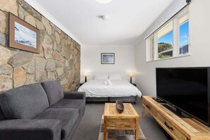 A cozy room with a stone wall, a bed with white bedding, a grey sofa, a wooden coffee table, and a TV on a wooden stand.