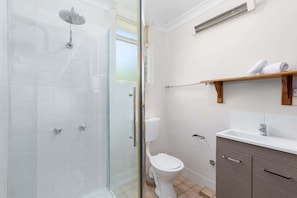 A bathroom with a glass-enclosed shower, a toilet, a sink, and a wooden shelf with towels.