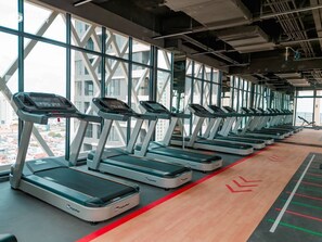 Varias caminadoras en un gimnasio con ventanas amplias y vistas a edificios.