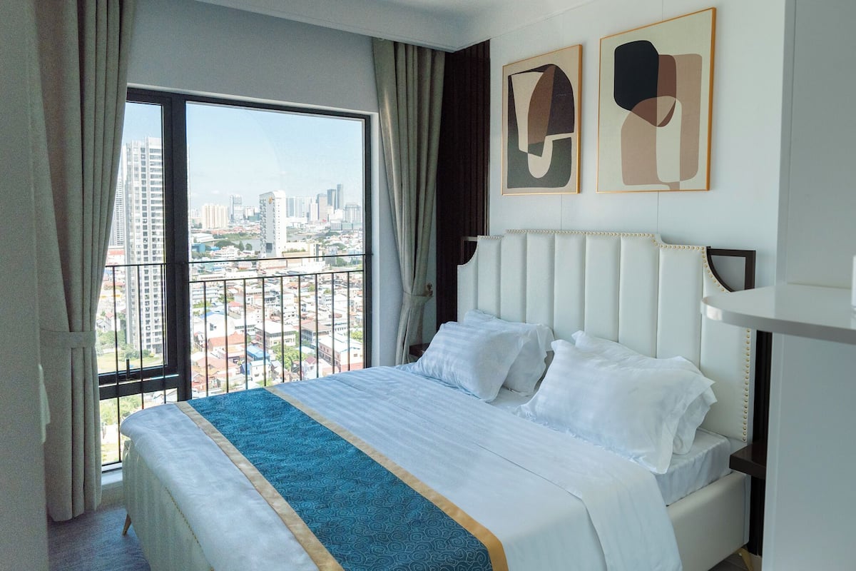 A hotel room with a bed, a ceiling fan, a city view, and abstract art on the wall.