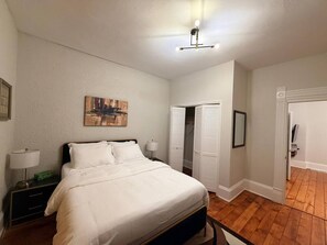 Ein Schlafzimmer mit Bett, Nachttischen, einem Gemälde, einem Kleiderschrank und einer Tür zu einem anderen Raum.