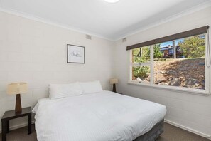 Ein Schlafzimmer mit Bett, zwei Nachttischen, einem Fenster mit Blick ins Freie und einem gerahmten Bild an der Wand.