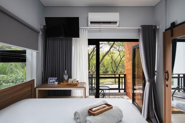 A hotel room with a bed, a bedside table with a tray, a TV, an air conditioning unit, and a balcony with a view of greenery.
