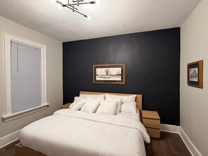Une chambre à coucher avec un lit, une table de chevet, un tableau encadré au mur et une fenêtre avec des stores.