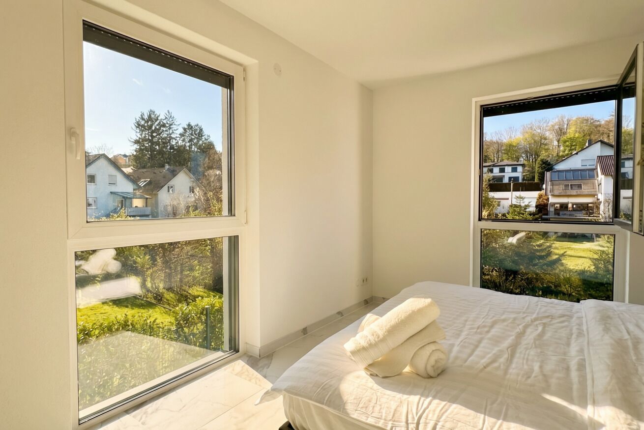 A bedroom with a bed, two windows, and a view of houses and greenery.