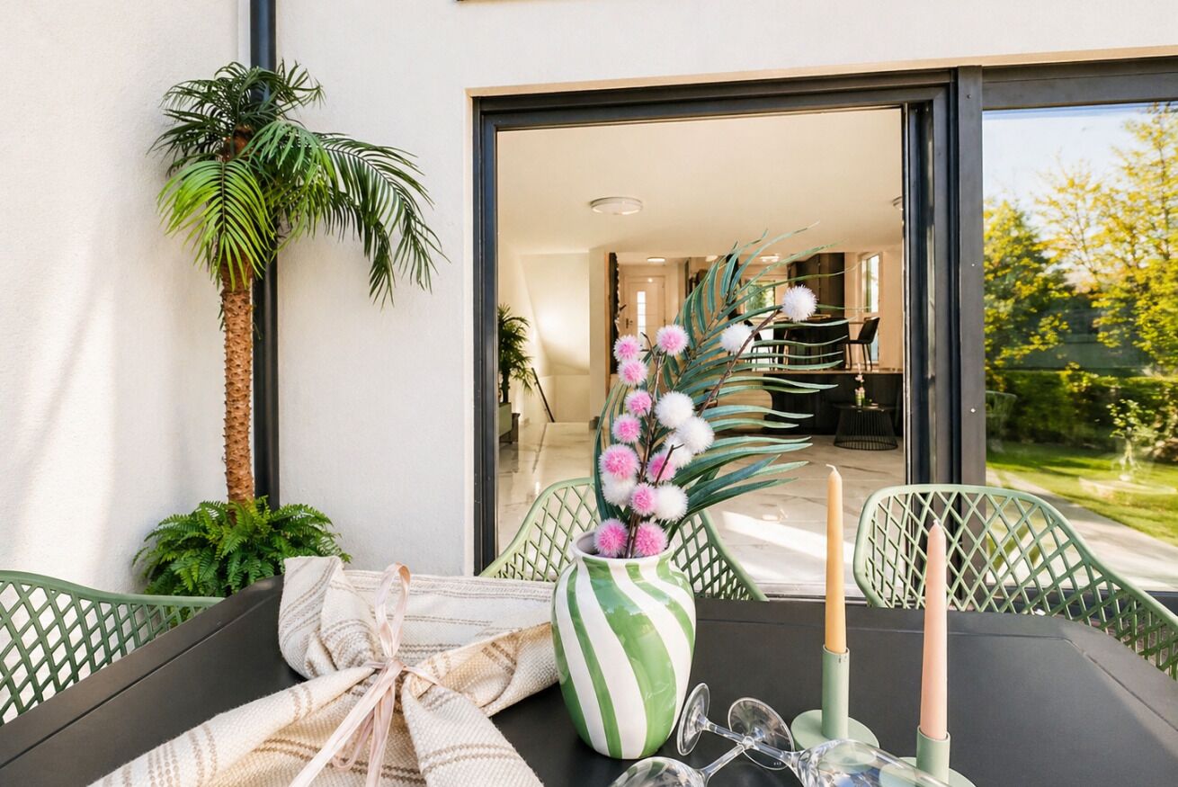 A dining area with a table set for two, a green chair, a striped vase with pink flowers, and a tall palm plant.
