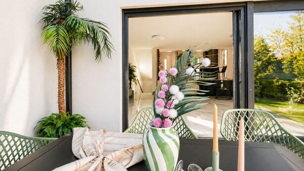 A dining area with a table set for two, a green chair, a striped vase with pink flowers, and a tall palm plant. - Elegant 4BR Villa w/ Garden & Terrace (Mödling)