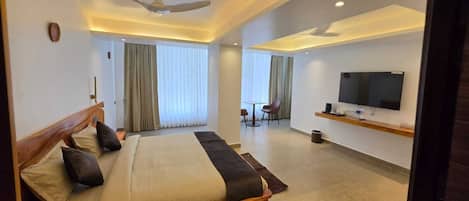 A modern hotel room with a large bed, a TV, a ceiling fan, and a dining area with a table and chairs.