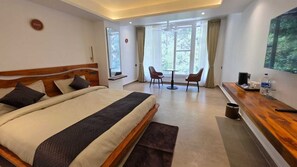 A modern hotel room with a large bed, a desk with a chair, a small table, and a large window. - Hotel Anukampa (Kosiyakutoli)