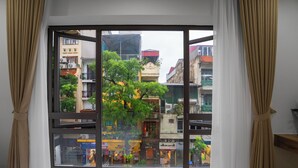 Ein Zimmer mit großem Fenster, Vorhängen, einem Bett und Blick auf eine Stadtlandschaft mit Gebäuden und Bäumen.