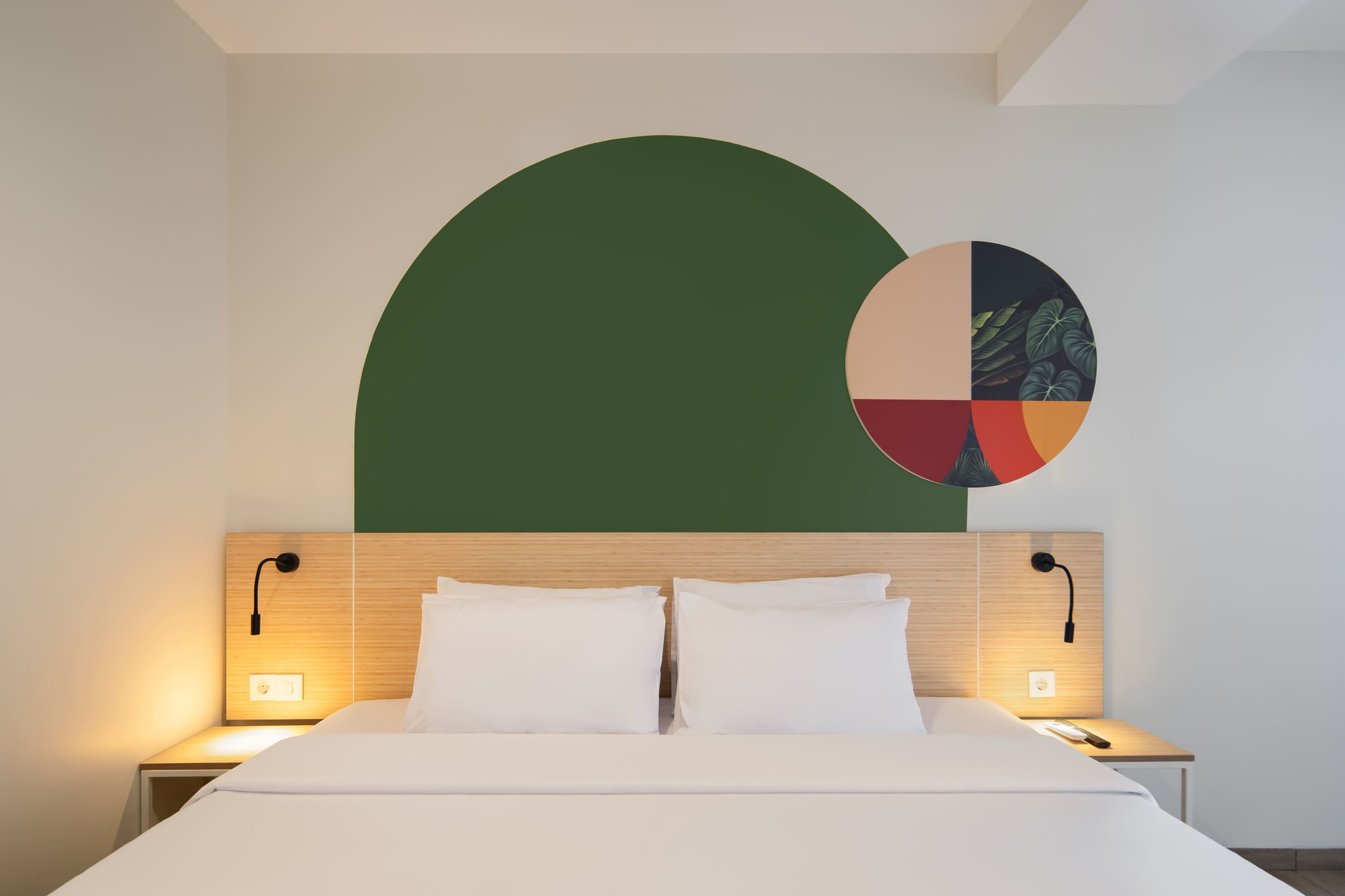 A hotel room with a bed, two bedside tables, a wall-mounted light, and a partial view of a large green and multicolored circular artwork.