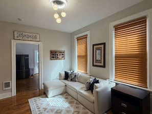Ein Wohnzimmer mit Ecksofa, Holzboden, einer Deckenleuchte und einer Küche im Hintergrund.