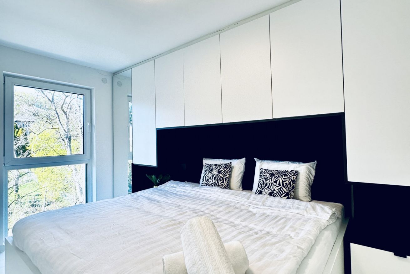 Un dormitorio moderno con una cama grande, paredes blancas y un ventanal con vistas a árboles.