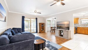 A living room with a sofa, TV, and cabinets, a dining area with chairs, and a kitchen with wooden cabinets. - Lynn Haven 3BR - Private Pool & Fenced Yard (Lynn Haven)