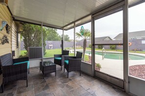 Ein überdachter Patio-Bereich mit Korbmöbeln, einem Glastisch und Blick auf einen Swimmingpool.