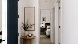 A hallway with a dark blue door, a round wooden table with a plant and candle, a framed picture on the wall, and a bedroom visible in the distance. - Brand-New Luxury Private ADU, 2BR/2BA, Full Kitchen & Laundry (Fountain Valley)
