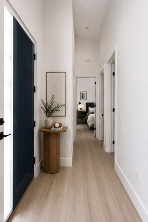 A hallway with a dark blue door, a round wooden table with a plant and candle, a framed picture on the wall, and a bedroom visible in the distance. - Brand-New Luxury Private ADU, 2BR/2BA, Full Kitchen & Laundry (Fountain Valley)