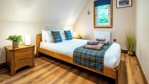 A bedroom with a wooden bed, a bedside table with a lamp, a window with a view of greenery, and a wall-mounted picture.