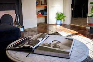 Un livre posé sur une table en marbre, dans une pièce où se trouvent une cheminée, une plante et une bibliothèque.