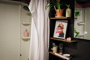 Une salle de bain avec un rideau de douche, un tableau encadré, une plante en pot, une petite étagère avec une bouteille rose et un miroir qui reflète une plante.