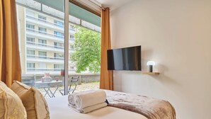 A hotel room with a bed, a TV mounted on the wall, and a window with a view of a building and trees. - Charming Suite With AC - Eiffel Tower-beaugrenelle (Paris)