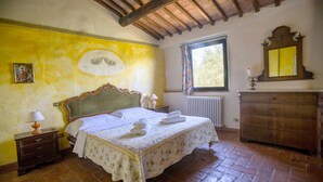 Ein Schlafzimmer mit Bett, Nachttisch, Kommode, Fenster mit Vorhängen und Heizkörper.
