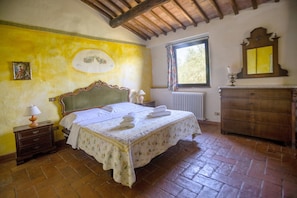 Ein Schlafzimmer mit Bett, Nachttisch, Kommode, Fenster mit Vorhängen und Heizkörper.