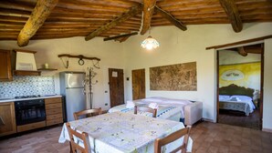 Eine rustikale Küche mit Herd, Kühlschrank und Holzschränken. Ein Esstisch mit Stühlen und ein Himmelbett sind im Hintergrund zu sehen.