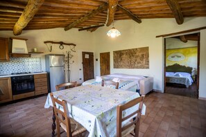 Eine rustikale Küche mit Herd, Kühlschrank und Holzschränken. Ein Esstisch mit Stühlen und ein Himmelbett sind im Hintergrund zu sehen.