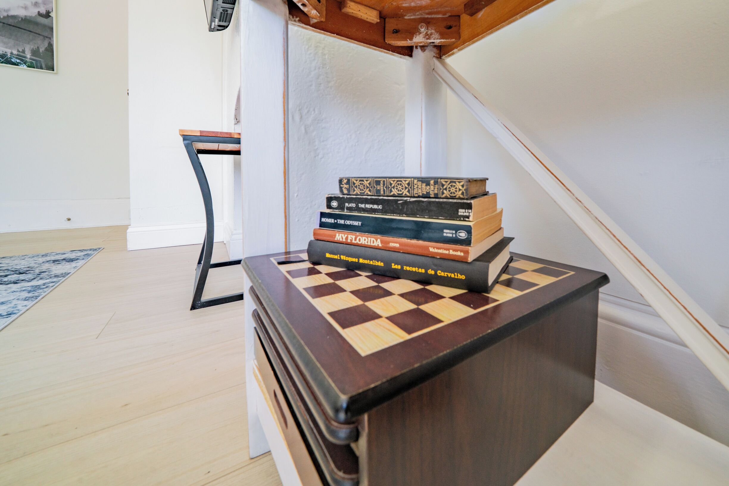 A wooden chest with a chessboard on top, stacked with books, in a room with a wooden floor and a white wall.