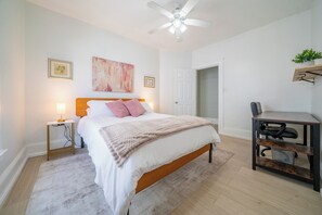 Ein Schlafzimmer mit einem Bett, Nachttischlampen, einem Deckenventilator, einem Schreibtisch mit Stuhl und einer Tür zu einem anderen Raum.