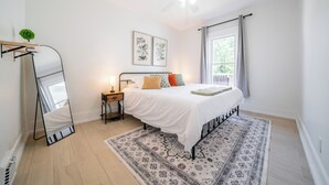 Ein Schlafzimmer mit einem Bett, einem Deckenventilator, einem Fenster mit Vorhängen, einem Nachttisch und einem Spiegel.