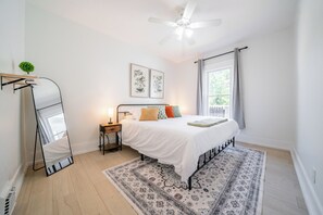 Un dormitorio con cama, ventilador de techo, ventana con cortinas, mesita de noche y espejo.