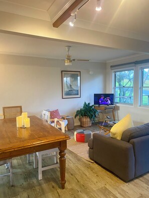 A living room with a wooden dining table, a sofa, a TV, and a ceiling fan. - “Southerly Change”.
Groovy retro/modern Beach House.
Bring your surfboard & dog! (Moruya Heads)